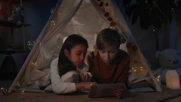 Подростки в декоративной импровизированной палатке с помощью планшета, разговаривая и улыбаясь вечером. Веселые мальчик и девочка лежат на полу, проводя свободное время. Концепция досуга и беспечности. — стоковое видео