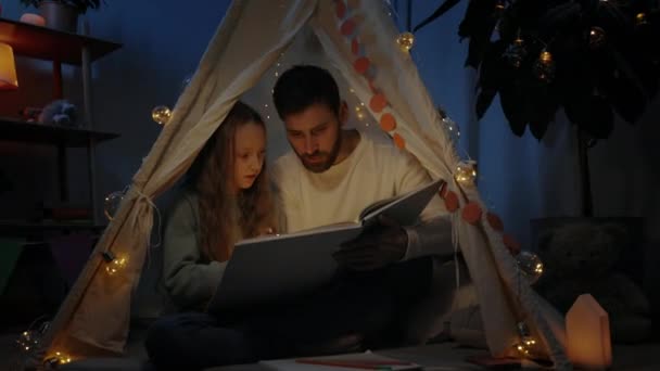 Pai feliz e sua menina adolescente lendo livro juntos e sorrindo enquanto sentado em cabana improvisada decorativa. Conceito de tempo familiar. — Vídeo de Stock