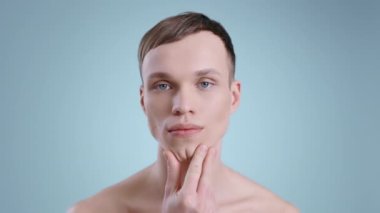 Milenyum insanının kameraya bakarken yüzüne dokunuşunun vesikalığı. Çıplak omuzları ve mavi gözleri olan yakışıklı bir erkek modelin portresi. Kozmetik ve cilt bakımı kavramı.