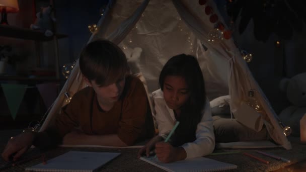 Ung dreng og pige taler og skaber billeder, mens du bruger fritid om aftenen. Teenagere liggende på gulvet og taler i dekorative improviseret hytte, mens tegning med farvede blyanter. – Stock-video