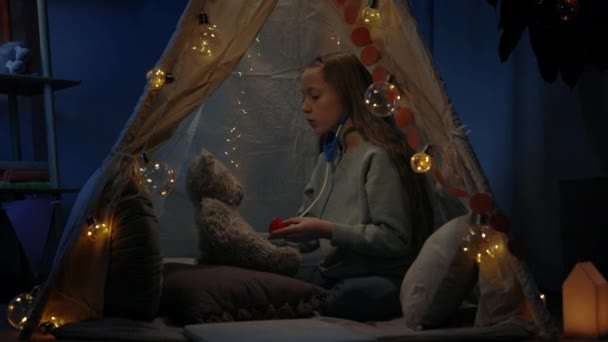 Ładna dziewczyna bawi się misiaczkiem siedząc na podłodze w dekoracyjnym prowizorycznym namiocie w domu. Dziecko z zabawkowym stetoskopem spędzające wolny czas wieczorem. Pojęcie czasu wolnego. — Wideo stockowe