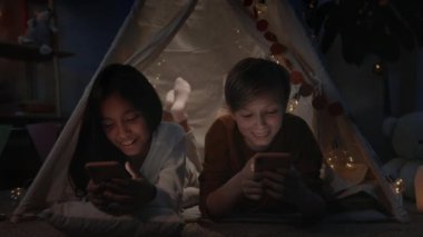 Neşeli gençler akşamları evlerinde dekoratif derme çatma kulübelerde internete göz atarken vakit geçiriyorlar. Kız ve erkek yerde yatarken ve akıllı telefon kullanırken gülüyorlar..