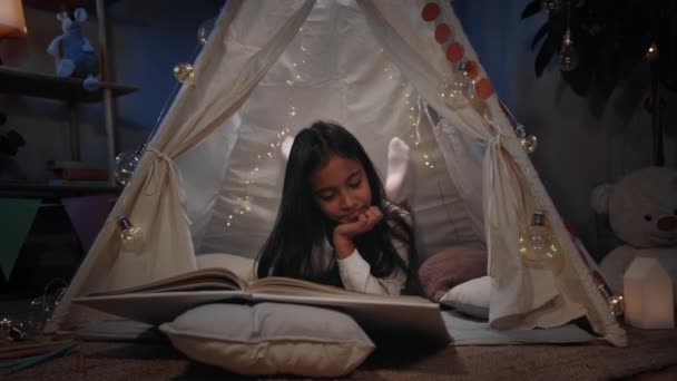 Дівчинка-підліток у красивій хатинці читає книгу вдома ввечері. Досить позитивний підліток лежить на підлозі, проводячи вільний час. Концепція дозвілля і недбале дитинство . — стокове відео