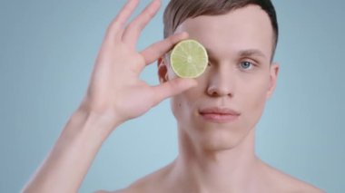 Kameraya bakarken limonun yarısını tutan yakışıklı adamın portresi. Çıplak omuzlu, açık mavi gözlü, meyveli genç bir erkek model. Cilt bakımı kavramı, sağlık sigortası..