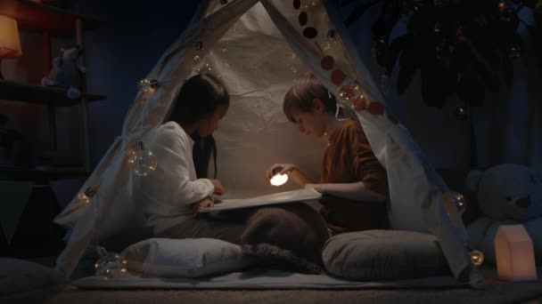 Tinédzserek dekoratív makeshift sátor keres és fordul a könyv oldalon este. Vidám fiú és lány zseblámpával és a földön ülve, miközben szabadidőt töltenek. A szabadidő fogalma. — Stock videók