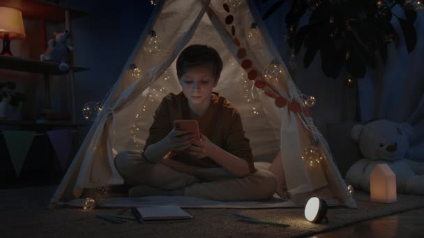 Pozitív fiú szőke hajjal böngészi az internetet, miközben este otthon dekoratív sátorban tölti az idejét. Férfi tini ül a padlón használata közben és görgetés okostelefon.koncepció a szabadidő. — Stock videók