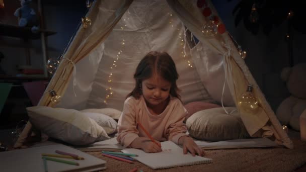 Cute little girl rysunek z kolorowych ołówków leżąc na podłodze w pięknym namiocie dekoracyjnym w domu. Ładny dzieciak spędza wolny czas wieczorem. Koncepcja dzieciństwa. — Wideo stockowe