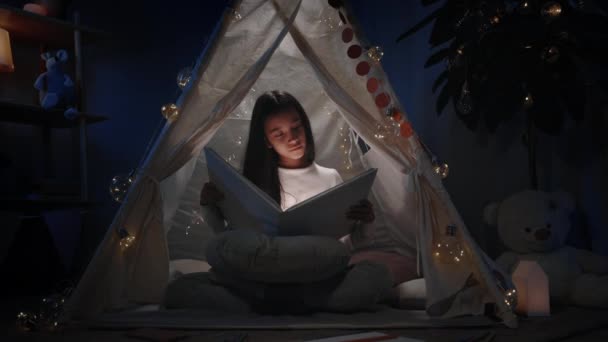 Menina inteligente com cabelo preto longo em tenda decorativa bonito olhando para reservar com lanterna. Muito adolescente sentado no chão enquanto lê. Conceito de lazer e tempo livre. — Vídeo de Stock