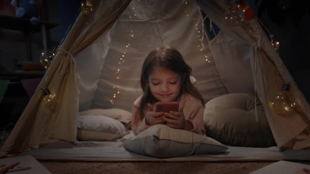 Adorable petite fille passant du temps dans une tente décorative avec des éclairages dans la salle de jeux du soir. Mignon petit enfant couché sur le sol et souriant tout en regardant le dessin animé sur smartphone. Zoom avant. — Video