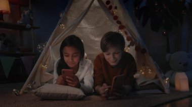 Delikanlı kız dekoratif çadırda yatarken ona akıllı telefon içeriğini gösteriyor. Gençler akşamları telefonlarını kullanıyor ve gülümsüyor. Boş zaman kavramı..