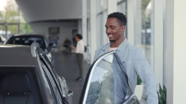 Hombre africano feliz de pie en la concesionaria cerca de coche de lujo — Vídeos de Stock