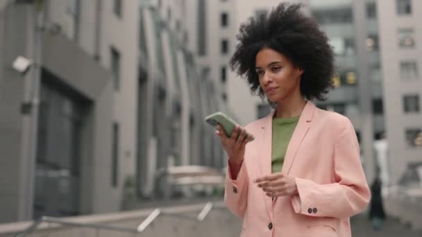 Donna multirazziale sorridente che indossa abito formale utilizzando il telefono cellulare e digitando messaggi di testo mentre cammina fuori dal centro business. Tecnologie e concetto di persone. — Video Stock