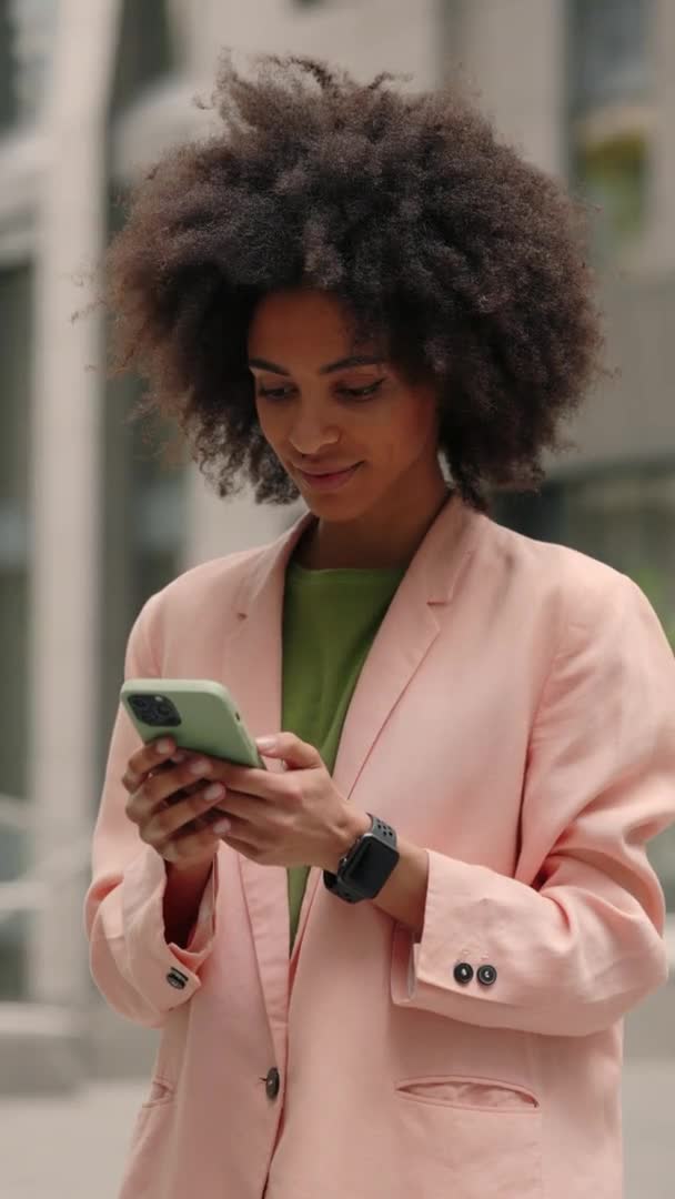 Vídeo vertical. Mujer joven multirracial sonriente en traje formal mirando a su teléfono inteligente fuera en la calle y regocijándose de algo. Gente de negocios emociones concepto. — Vídeo de stock