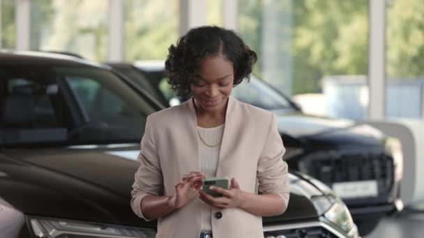 Mujer africana sonriente utilizando el teléfono inteligente al elegir el coche — Vídeo de stock