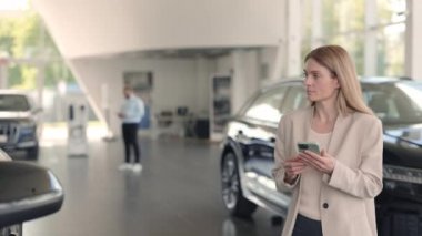 Kadın elinde akıllı telefonuyla araba galerisinde yürüyor.