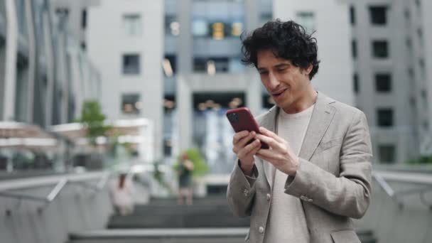 Усміхнений кавказький молодий чоловік в офіційному вбранні дивиться на свій смартфон на вулиці і радіє чомусь. Бізнес сучасний менеджер і концепція емоцій людей . — стокове відео