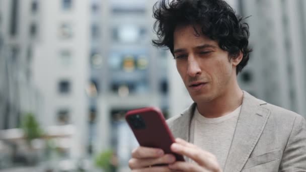 Hombre de negocios caucásico en un traje está utilizando un teléfono inteligente en una calle en el centro de la ciudad mientras navega por la web en su dispositivo. Sonríe y parece exitoso. Concepto de personas y tecnologías. — Vídeo de stock