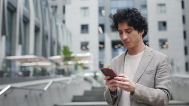 Başarı. Şehir merkezindeki caddede duran iş adamının kamera görüntüsünü değiştir ve telefonuyla mesajlaşırken, kaydırırken ve gülümseyerek tıkırdatırken. Teknoloji ve iletişim konsepti.