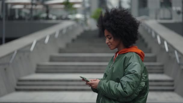 Per strada. Vita in su ritratto vista della donna multirazziale calma con i capelli ricci in piedi per strada e guardando lo schermo del suo smartphone. Tecnologie e concetto di persone. — Video Stock