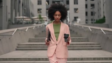 Elinin altında belgelerle ve akıllı telefonuyla iş binaları arasında yürüyen ve neşeyle gülümseyen başarılı genç ve çok kültürlü bir kadın. Çekici iş kızı ve kariyer insanı konsepti.
