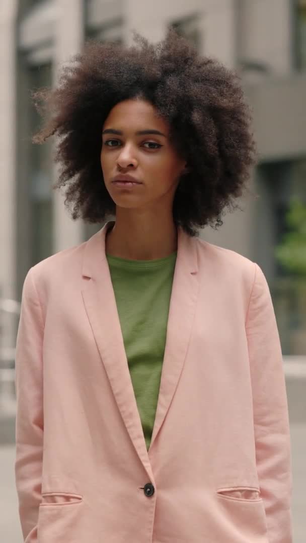 Vídeo vertical. Retrato de la mujer de negocios con el pelo rizado mirando a la cámara mientras se para en la ciudad de la calle y compartir emociones graves. Estilo de vida de verano chica ciudad al aire libre concepto. — Vídeos de Stock