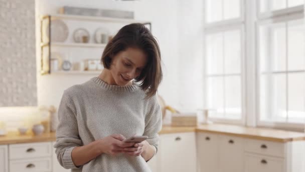 Genç bir kadın mutfaktayken modern akıllı telefon kullanıyor. Kafkasyalı esmer kadın evde boş zamanlarında gezerken internette sörf yapıyor.. — Stok video