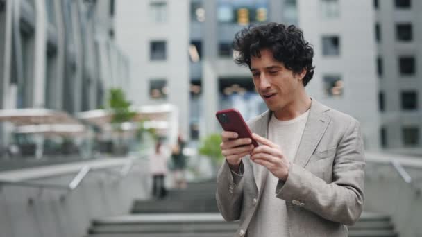 L'uomo d'affari caucasico in giacca e cravatta sta usando uno smartphone in una strada in centro mentre naviga sul web sul suo dispositivo. Sorride e sembra di successo. Concetto di persone e tecnologie. — Video Stock