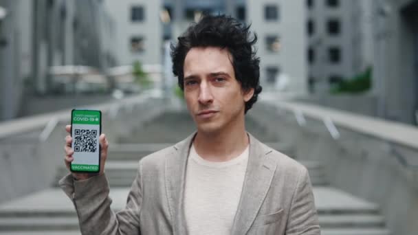 Retrato del hombre de negocios mostrando su smartphone con pasaporte de vacuna de pie cerca del centro de negocios. — Vídeos de Stock