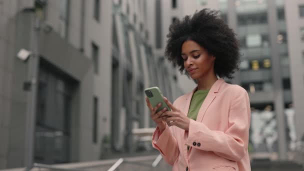 Usmívající se multiraciální žena ve formálním obleku pomocí mobilního telefonu a psaní textových zpráv při chůzi mimo obchodní centrum. Koncept technologií a lidí. — Stock video