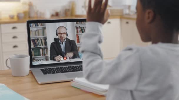 View from shoulder of pretty child greeting male teacher thought video call on modern laptop. African school boy having online education during quarantine. — Wideo stockowe