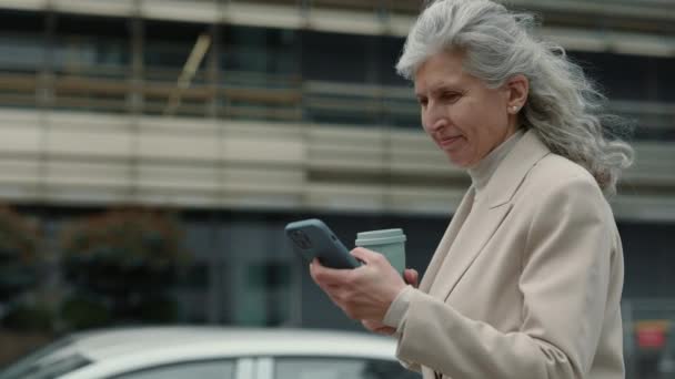 Пожилая деловая женщина с кофе и мобильной ходьбы по улице — стоковое видео