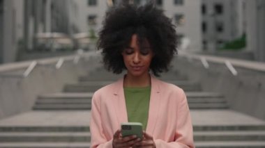 Modern insanlar. Çok ırklı kıvırcık kadının akıllı telefonunu kullanırken ve şehirde poz verirken kameraya bakarken hareketli kamera görüntüsü.