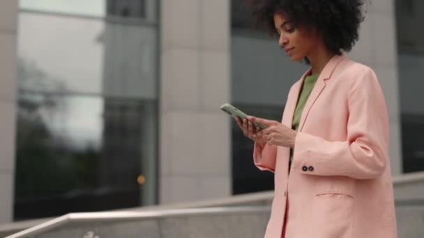 Beautiful young businesswoman wearing formal suit and using modern smart phone while walking at break in the city. Professional female employer typing text message on cellphone outside. — Stock Video