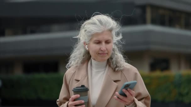 Пожилая деловая женщина в наушниках с помощью мобильного телефона на улице — стоковое видео