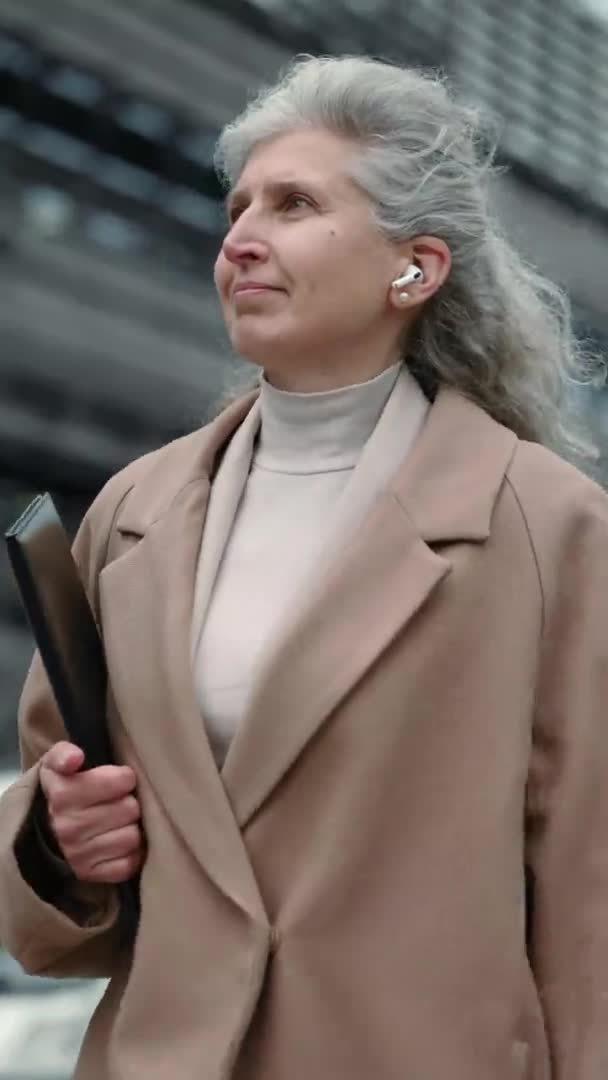 Vidéo verticale. Femme âgée dans les écouteurs sans fil portant presse-papiers à l'extérieur — Video