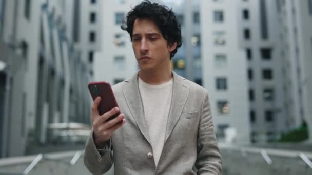 Retrato del hombre de negocios sorprendido mirando teléfono inteligente fuera y caminando cerca del centro de negocios. Retrato del hombre emocionado leyendo buenas noticias en el teléfono móvil al aire libre. Hombre sintiéndose feliz y emocionado. — Vídeo de stock