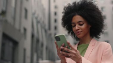Karışık ırklı kıvırcık kadının cep telefonuyla bir şeylere göz atarken ve sokaklarda kentsel binalarla vakit geçirirken keyif gülümsemesi gösterirken düşük açılı görüntüsü. Teknoloji konsepti.