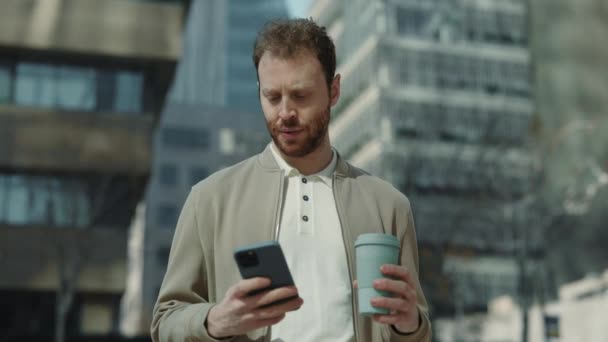 Hombre joven con café y teléfono inteligente de pie al aire libre — Vídeos de Stock