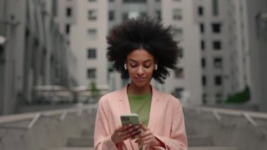 Enjoying of the sound. Waist up portrait view of the curly multiracial woman wearing earphones listening music and feeling great while spending her day at the street.