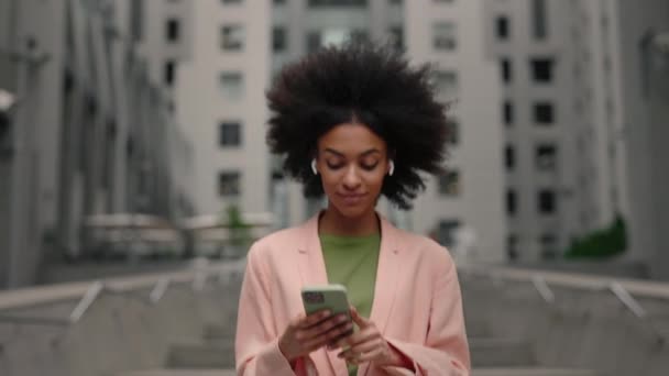 Enjoying of the sound. Waist up portrait view of the curly multiracial woman wearing earphones listening music and feeling great while spending her day at the street. — ストック動画