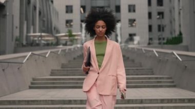 Successful young businesswoman with documents under her hand and smartphone walking between business buildings and smiling with joy. Attractive business girl and career people concept.