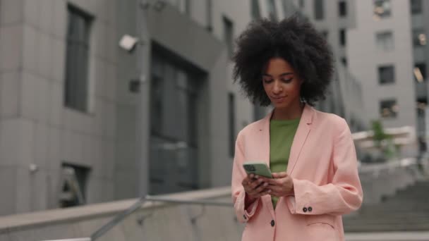 Vista de retrato de la mujer de negocios rizada mixta navegando algo en su teléfono móvil y mostrando una sonrisa de placer mientras pasa tiempo en la calle con edificios urbanos. Concepto de tecnologías. — Vídeos de Stock