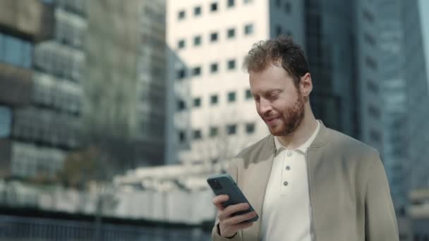 Позитивный бородатый мужчина ходит по улице с мобильным телефоном в руках — стоковое видео