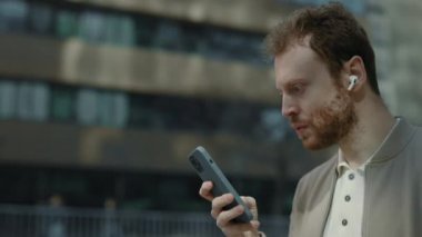 Şaşkın kulaklıklı adam sokakta cep telefonuyla yürüyor.