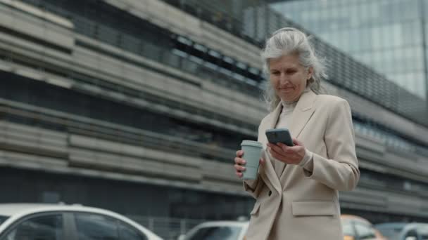 Gammal kvinna med mobil och kaffe promenader på gatan — Stockvideo