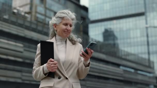 Старшая женщина с мобильным телефоном и планшетом ходит по улице — стоковое видео