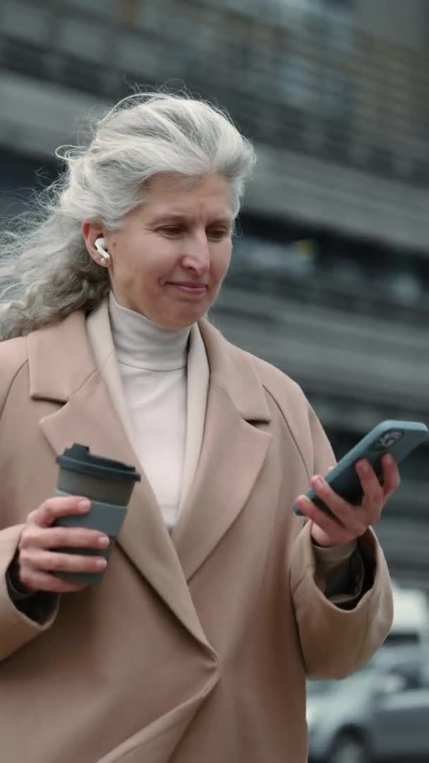 Dikey video. Kulaklıklı yaşlı bayan sokakta yürüyor ve cep telefonu kullanıyor. — Stok video