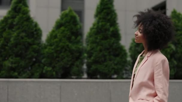 Cámara de movimiento vista de la feliz mujer de negocios multirracial que lleva traje formal caminando por la calle de verano y saludando a alguien mientras está de buen humor. — Vídeos de Stock