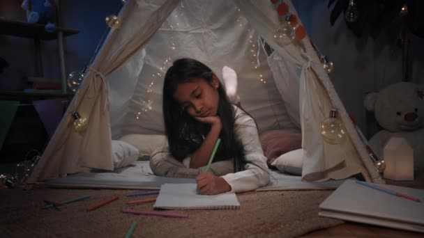Söt flicka med långt svart hår ritning med färgade pennor när du ligger på golvet i dekorativa tält hemma. En söt kvinnlig tonåring som tillbringar ledig tid på kvällen. Begreppet kreativitet. — Stockvideo