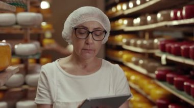 Erkek teknoloji uzmanı peynir kafalarının durumunu incelerken, kadın süt ürünleri müfettişi dijital tablet üzerinde çalışıyor. Lezzetli organik yiyecekler üreten iki uzman..